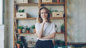 Woman thinking, contemplating, hesitating standing in front of a tidy shelf filled with plants and decorative boxes